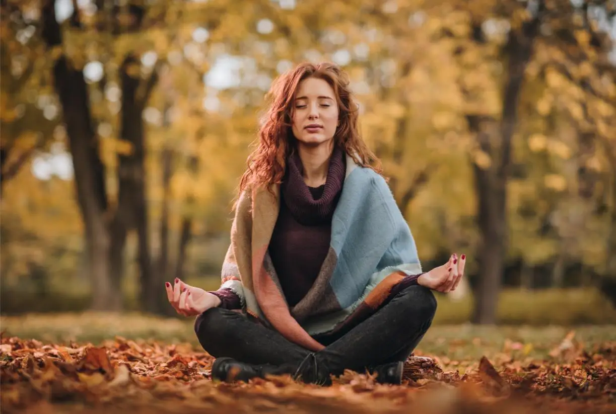 Ginger woman doing Yoga breathing exercises with her eyes closed during autumn day.